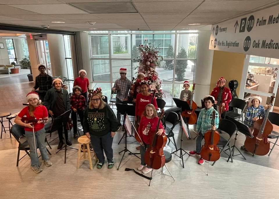 Students in Santa hats playing violins, cellos and a double bass beside a Christmas tree in a busy lobby.