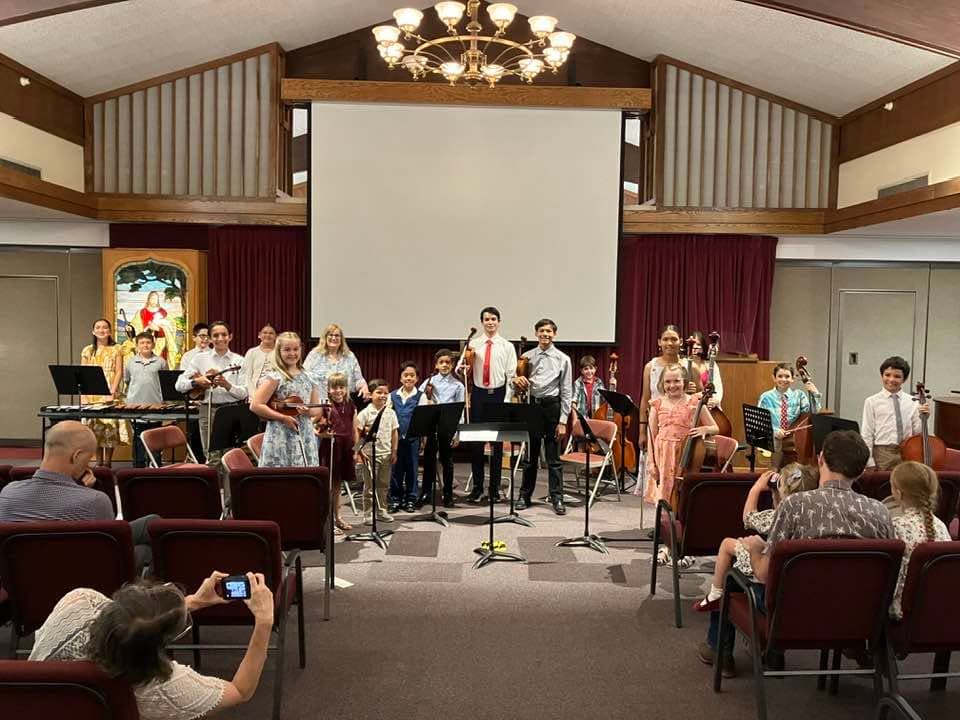 A large group of students standing on stage with their instruments at the end of a recital, families watching from the seats.