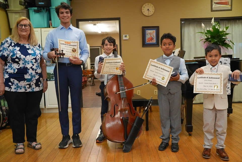 Four students holding certificates of achievement after a recital, one with a double bass, beside their teacher.