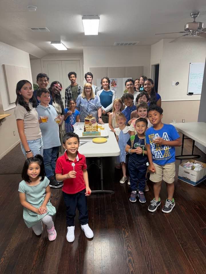 Students of all ages gathered around a cake decorated for a composer birthday celebration.