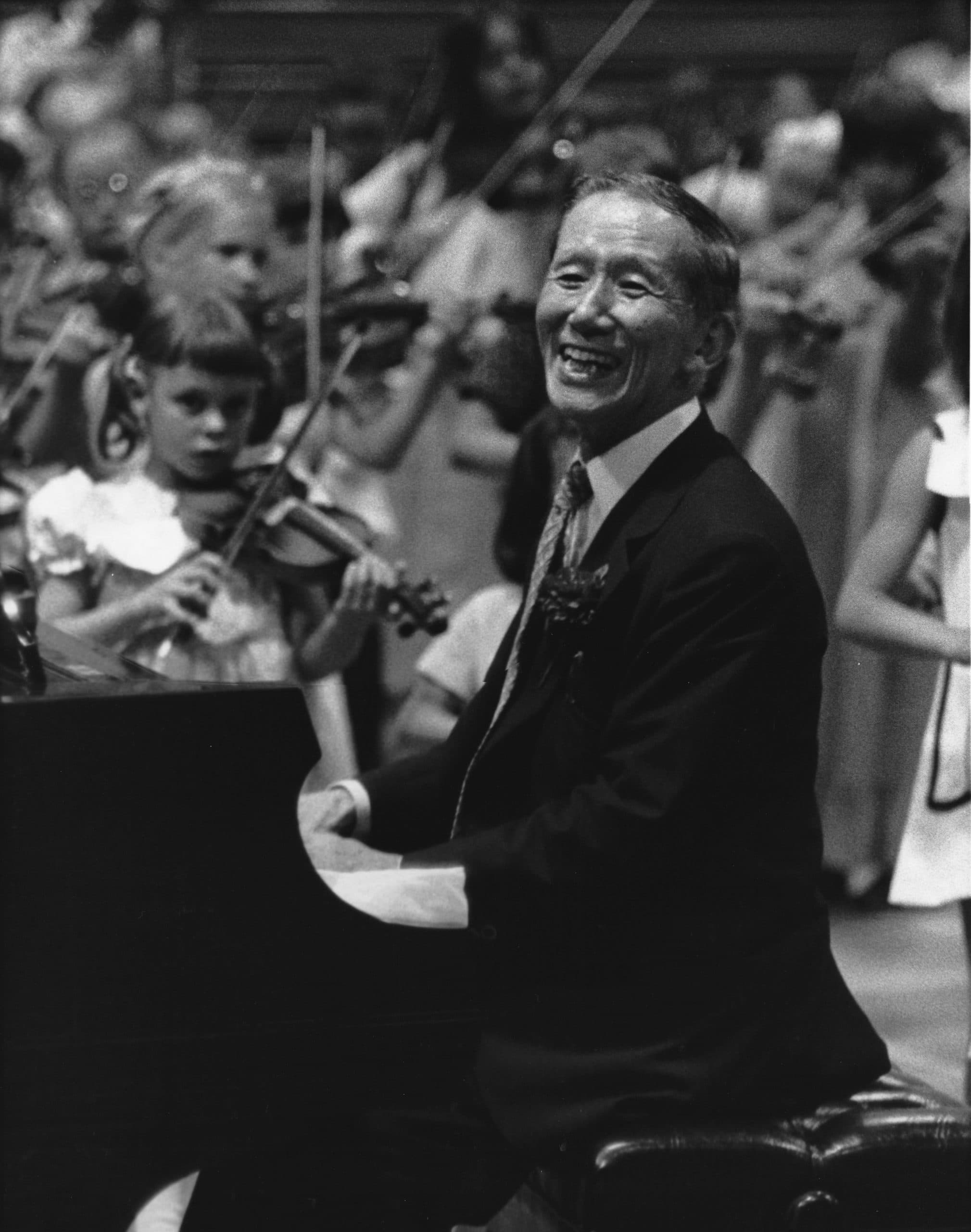 Dr. Shinichi Suzuki seated at a piano, smiling, with a group of young violin students playing behind him.